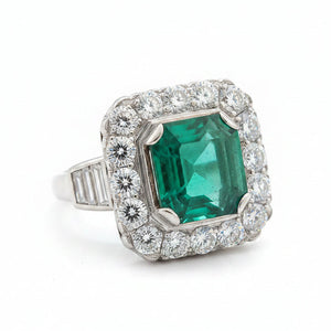 A platinum ring featuring a square-cut green emerald gemstone surrounded by a halo of clear diamonds, with baguette-cut side stones on the band.
